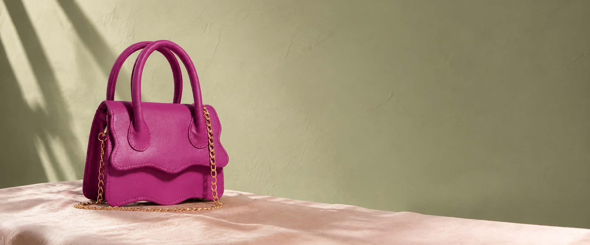 Magenta Muffin handbag with scalloped edges and a gold chain on rose linen against a sunlit sage wall.